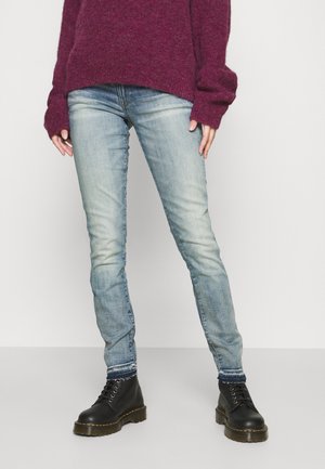 Personne portant un jean skinny bleu clair délavé, des bottes noires à lacets et un pull bordeaux à manches longues, debout devant un fond uni.