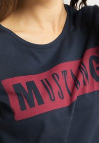 Navy blue cotton t-shirt featuring a burgundy horizontal band with large black text reading "MUSTANG." Crew neck design, soft texture.