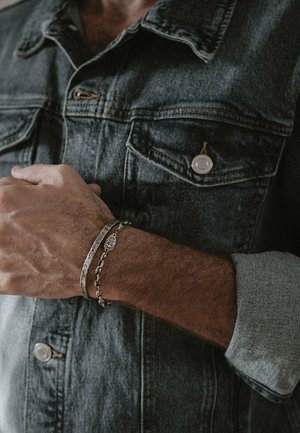 Silver bracelets on a wrist, layered. One features intricate detailing; the other has chain links. Hand rests against a denim jacket.