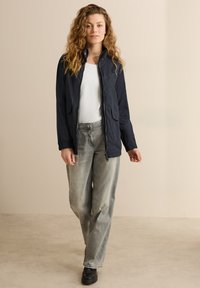 Marineblauwe softshell jas met een ritssluiting aan de voorkant, twee zijzakken, en elastische manchetjes, gedragen over een wit shirt, gecombineerd met lichtgrijze wijde jeans.