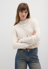 Roomkleurige kabelgebreide cardigan met een ronde halslijn, geribbelde manchetten en knopen aan de voorkant, gedragen over een wit kanten topje, gecombineerd met blauwe spijkerbroeken.