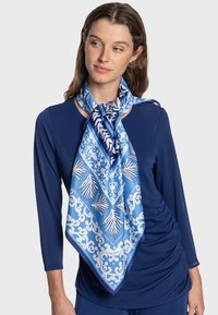 Écharpe en soie bleue ornée de motifs floraux et géométriques blancs complexes, drapée élégamment autour du cou, complétant un haut bleu marine.