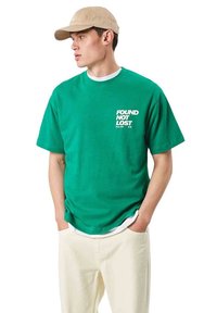 Joven con gorra beige, camiseta verde con el texto "FOUND NOT LOST", camiseta blanca debajo y pantalones color crema, con las manos en los bolsillos.