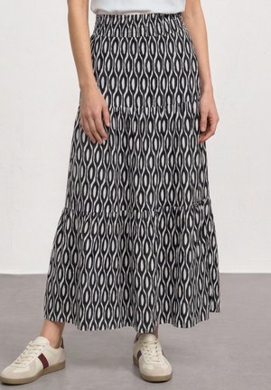 Person iført et sort og hvidt geometrisk mønstret maxinederdel, hvid top og hvide sneakers med brune detaljer, står indendørs.