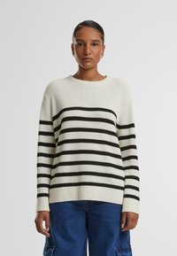 Maglione bianco lavorato a maglia con righe orizzontali nere, polsini a coste e scollo rotondo. Abbinato a pantaloni in denim blu larghi.