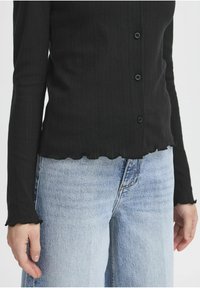 Černý žebrovaný kardigan s vlnkovaným lemem a manžetami, s pěti černými knoflíky, spárovaný se světle modrými džíny.