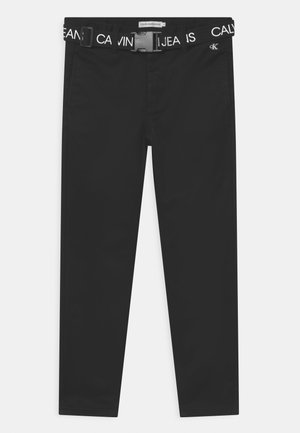 Pantalon noir à texture lisse, doté d'une ceinture blanche de marque visible avec l'inscription "CALVIN KLEIN JEANS" et une boucle en plastique.