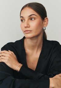 Egy fekete blúzt viselő személyt díszítenek ezüst karika fülbevalók és egy finom ezüst karkötő, aki hosszú, egyenes hajú, semleges háttér előtt áll.