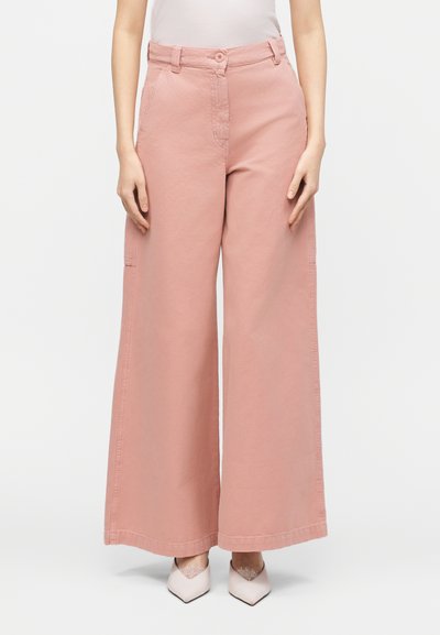 Femme portant un pantalon rose taille haute à jambes larges avec des poches avant et des chaussures blanches à bout pointu, debout devant un fond blanc uni.