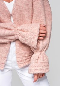 Světle růžový pletený kardigan s oversized zvonovitými rukávy, texturovanými detaily a komfortním designem s otevřeným předním dílem. Nositý přes bílý top.