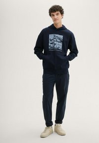 Granatowy hoodie z graficznym nadrukiem i tekstem, w połączeniu z dopasowanymi granatowymi spodniami i jasnymi trampkami. Miękki materiał i luźny krój.