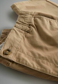 Beige Baumwollshorts mit glatter Textur, ausgestattet mit einem Knopfverschluss, gefälteltem Bund und einem teilweise sichtbar an der Naht angebrachten Etikett.