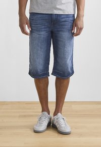 Pantalones cortos de mezclilla en azul medio, ligeramente desgastados, con corte a la altura de la rodilla y dobladillo enrollado. Combinados con zapatillas atléticas de color gris claro.