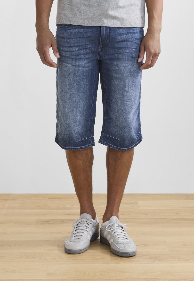Pantalones cortos de mezclilla en azul medio, ligeramente desgastados, con corte a la altura de la rodilla y dobladillo enrollado. Combinados con zapatillas atléticas de color gris claro.