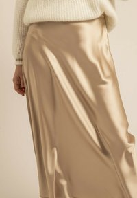 Jupe midi en satin beige clair, présentant une texture lisse et un design fluide, partiellement visible avec un haut en maille et un détail de main.