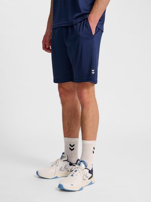 Mann trägt marineblaue Sportshorts, weiße Kniestrümpfe mit schwarzen Chevronen und weiße Turnschuhe mit blauen und schwarzen Akzenten, stehend.
