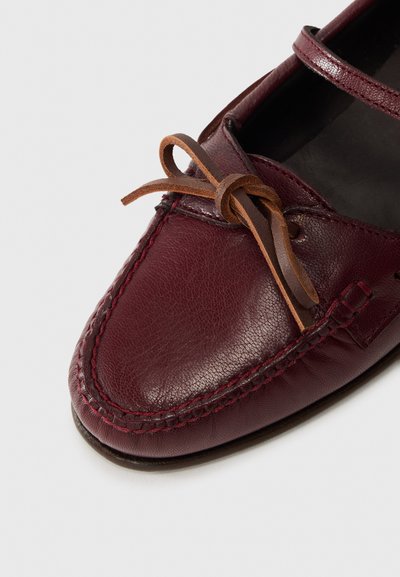 Zapato de cuero burdeos con punta redondeada, que presenta costuras tonales, una cinta delgada y un acento en color marrón. Textura suave con un diseño minimalista.