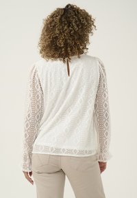 White lace blouse with geometric patterns, keyhole back detail, long sleeves, and decorative hem. Paired with light beige pants.