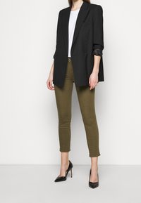 Blazer negro de doble botonadura sobre una blusa blanca, combinado con pantalones ajustados verde oliva y tacones negros de punta. Tela suave.