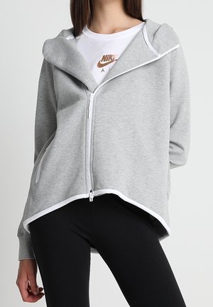 Kvinde iført en grå lynlås-hættetrøje med hvide detaljer over en hvid Nike t-shirt og sorte leggings, stående med den ene hånd afslappet ned langs siden.