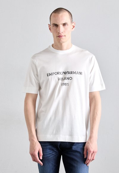 Fehér pamut póló középen fekete logónyomattal, melyen az "EMPORIO ARMANI MILANO 1981" felirat olvasható, kerek nyakkivágással és rövid ujjal.