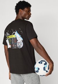 Camiseta preta com bilhetes de viagem impressos coloridos e ícones nas costas. O modelo segura uma bola de futebol com padrão branco e azul.