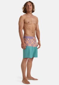 Pánske plavky s geometrickým vzorom v purpurovej a broskyňovej farbe v hornej časti, prechádzajúce do jednoliatej tealovej látky v dolnej časti.