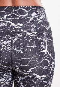 Leggings noirs avec un motif marbré blanc, dotés d'une large taille et d'un tissu texturé. Logo du fabricant visible près de la taille.
