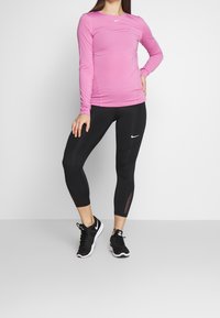 Pitkähihainen vaaleanpunainen urheilupaita ja mustat capri-legginssit, joissa on verkkomaisia yksityiskohtia; molemmissa on Nike-logot. Mustat urheilukengät täydentävät asukokonaisuutta.