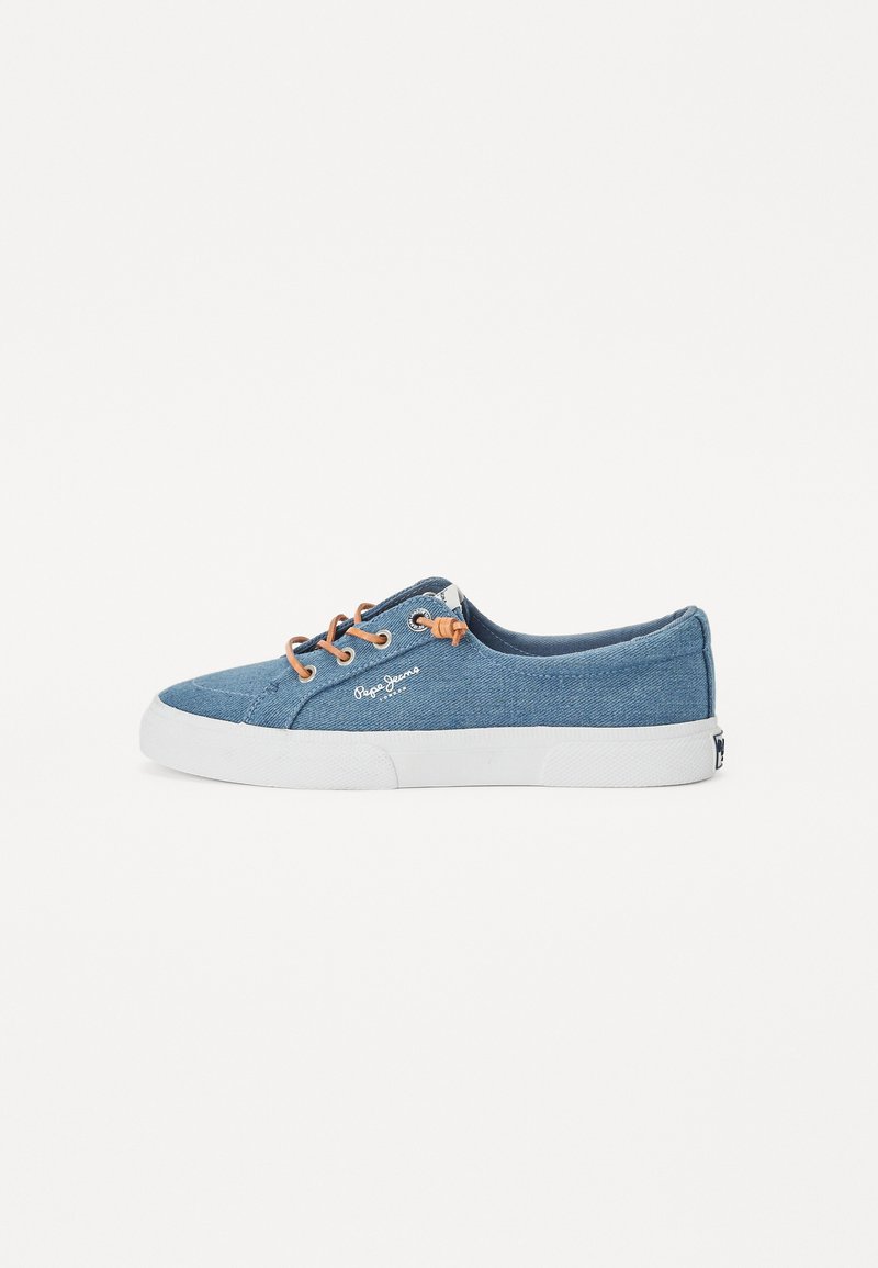Sapatilha de cano baixo em denim azul com sola de borracha branca. Apresenta atacadores castanhos e detalhes cosidos. Logótipo visível na lateral.