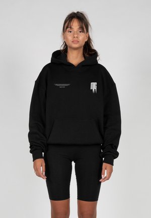 Junge Frau mit dunklem Pferdeschwanz trägt einen schwarzen, übergroßen Hoodie mit weißem Text und schwarzen Radlerhosen, stehend vor einem schlichten Hintergrund.