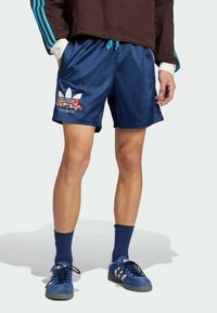 Marina atletiska shorts med resårmidja, blå dragsko, sidofickor och blommig Adidas-logotyp, i kombination med blå mockaskor och strumpor.