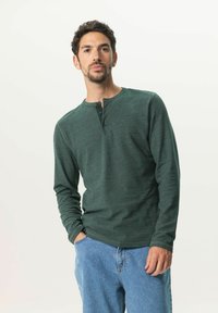 Chemise henley à manches longues verte en tissu texturé, avec un col rond et une patte de boutonnage à trois boutons, assortie à un jean en denim clair.