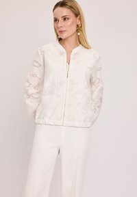 Biała kurtka zapinana na zamek z fakturowanym wzorem, elastycznymi mankietami i dołem, wykonana z lekkiego materiału, w zestawie z dopasowanymi białymi spodniami.