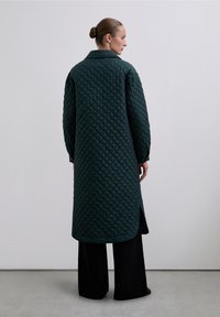 Abrigo largo acolchado en verde oscuro, con cuello, aberturas laterales y patrón de diamante texturizado. Lleva puesto sobre pantalones negros.
