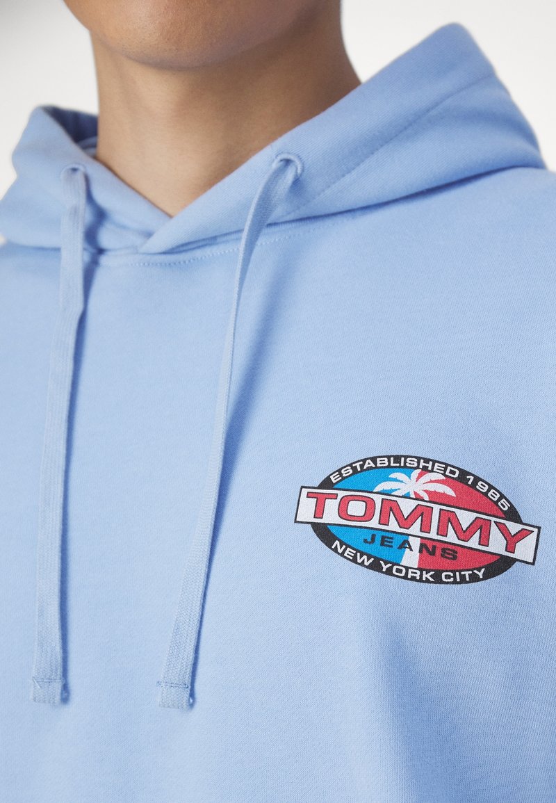 Gros plan d'un sweat à capuche bleu clair avec un logo Tommy Jeans représentant un palmier et le texte « Established 1985, New York City ».