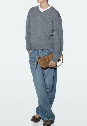 Persoon draagt een grijze trui en blauwe spijkerbroek, houdt een bruine suède crossbodytas vast, met één hand in de spijkerbroekzak.