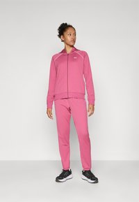 Roza zip-up atletska jakna s pripadajućim hlačama. Izrađena od mekanog materijala, s logom i kontrastnim bijelim šavovima. Uparena s crnim sportskim cipelama.