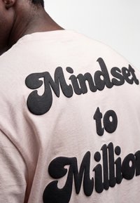 Persona che indossa una camicia rosa chiaro con testo nero in rilievo con scritto "Mindset to Millions" sulla schiena.