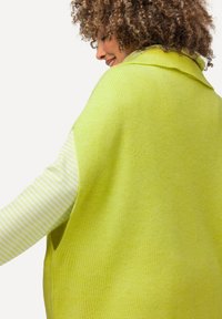 Maglione oversize verde lime con una finitura testurizzata e colletto ripiegato, abbinato a una camicia a maniche lunghe bianca e verde a righe sottostante.