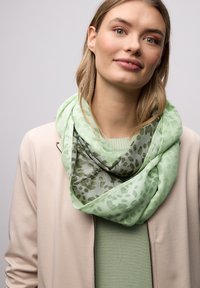 Femme aux cheveux châtain clair portant une veste beige par-dessus un haut vert et une écharpe légère à motifs verts autour du cou, sur un fond uni.