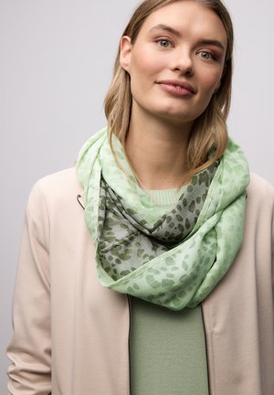 Femme aux cheveux châtain clair portant une veste beige par-dessus un haut vert et une écharpe légère à motifs verts autour du cou, sur un fond uni.