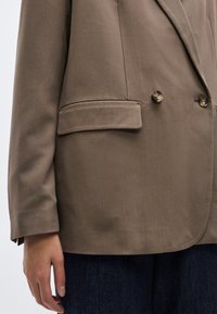 Καφέ διπλόμπρεστο σακάκι με απαλή υφή, δύο κουμπιά και μία θήκη στο στήθος. Τα μανίκια έχουν απλό μανσέτι.