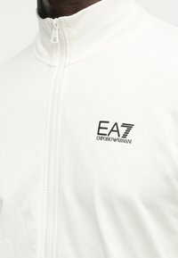 Κοντινό πλάνο ενός ατόμου που φοράει ένα λευκό μπουφάν με φερμουάρ, υψηλό γιακά και μαύρο λογότυπο EA7 Emporio Armani στο στήθος.