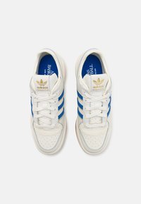 Valkoiset tennarit, joissa on sinisiä ja kultaisia yksityiskohtia, teksturoitu nahkapinta, rei'itetty varvasosa, nauhat ja kumipohja.