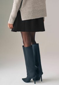Schwarze Leder-Stiefel bis zum Oberschenkel mit spitzen Zehen und Stiletto-Absätzen, kombiniert mit einem schwarzen Rock und einem grauen Strickpullover.