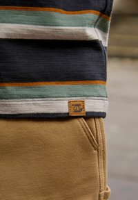 Stribet langærmet skjorte i navy, grøn, hvid og orange farver, med et vævet mærke; parret med tan bukser med lomme detalje.