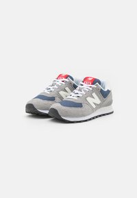 Zapatillas de running New Balance 574 en gris y marino con parte superior de malla y ante, logo blanco, lengüeta roja y blanca, y suela exterior de goma negra.