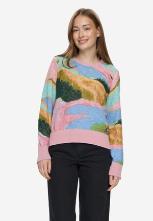 Mladá žena v barevném svetru s krajinným vzorem, růžovými manžetami a límcem, v černých kalhotách, usmívající se do objektivu.