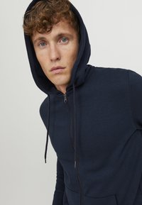 Marineblå zip-up hættetrøje med en frontlomme, snøre og blødt stof. Hætten er større og indrammer ansigtet.
